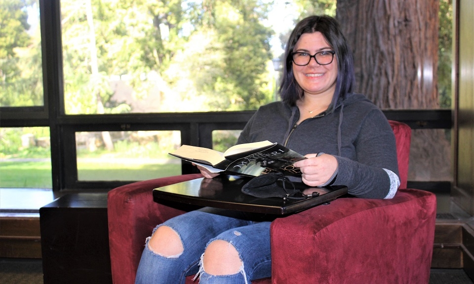 New photo of Abby Gordon smiling while sitting in chair book in hands in Alemany Library with redwood tree outside window over her left shoulder