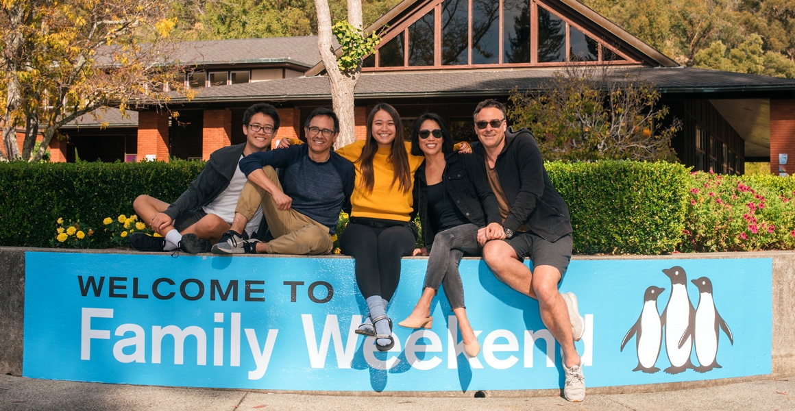 A family smiling at the camera outside of Caleruega Dining Hall. They are sitting on a low cement wall wrapped with a blue  banner that reads Welcome to Family Weekend.