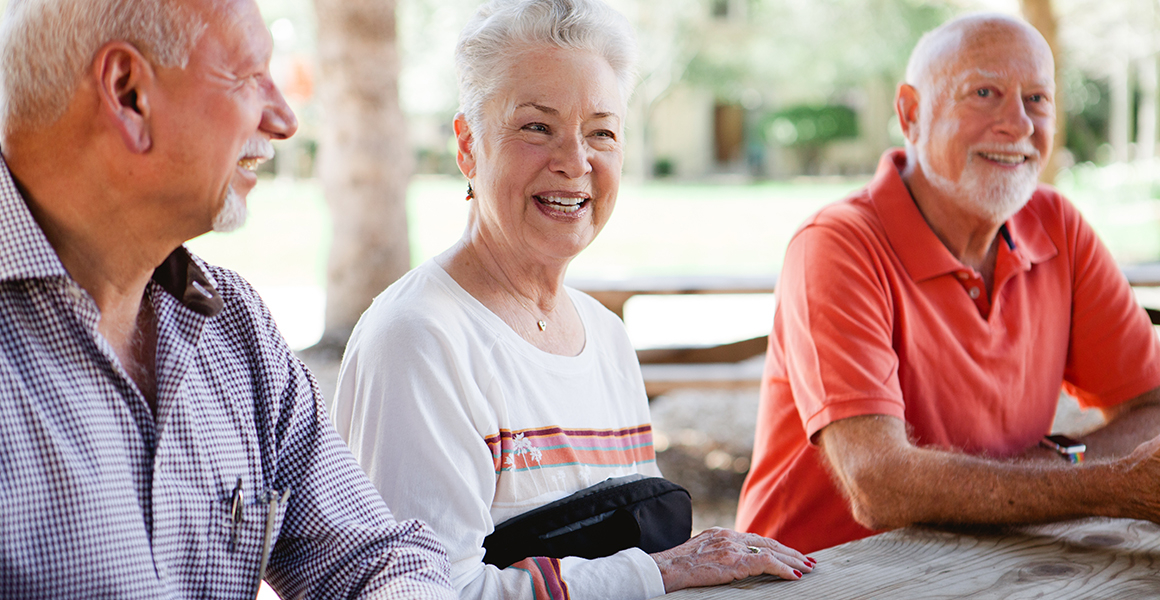 Three older adults sitting at an outdoor picnic table, smiling and talking.