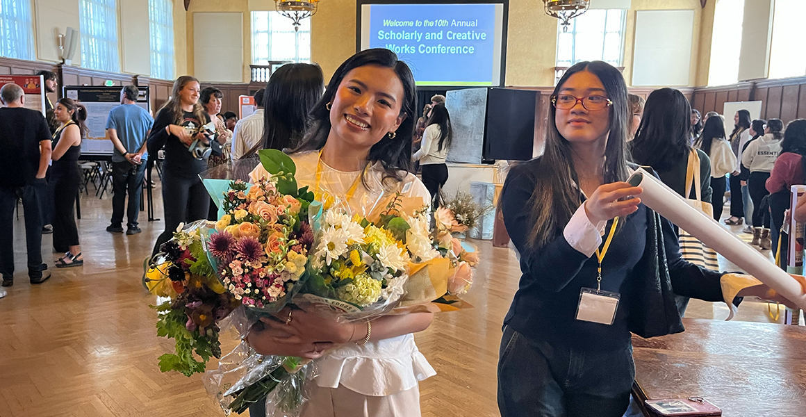 student with flowers in front of a sign with the scholarly and creative works conference on it