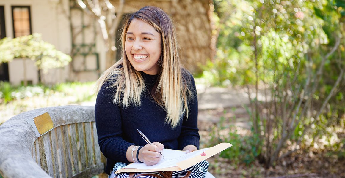 A young smiling woman is sitting on a wooden bench outdoors on the Dominican campus.