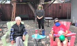 Three women sit and stand outside in a backyard during the Covid pandemic; two are wearing face masks. 