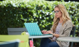 young woman with glasses sits alone typing on her laptop at an outdoor table