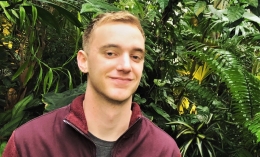 Photo of Alister Greenfield '17 wearing maroon jacket with green shrub background for HP image