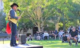 Photo of Sky Road Webb of Coast Miwok Council of Marin on stage speaking to audience on Indigeneous Peoples' Day on Anne Hathaway Lawn with Guzman Hall in background