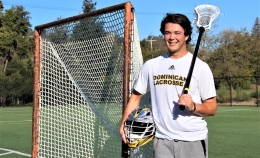 Photo of MBA student Maxwell Pierce wearing white Dominican lacrosse t-shirt standing next to lacrosse goal holding helmet with right hand and lacrosse stick over his left shoulder