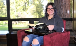 New photo of Abby Gordon smiling while sitting in chair book in hands in Alemany Library with redwood tree outside window over her left shoulder