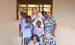 Photo of student-athlete Jordan Charlton standing wearing white Dominican Athletics long-sleeved T-shirt posing and smiling with arms around young Ugandan students smiling and wearing gray Dominican athletics T-shirts