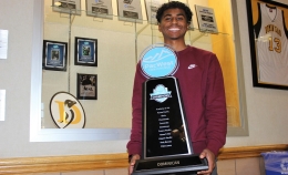 Photo of Maxwell Ward with maroon top standing and smiling in front of Conlan Center trophy case while holding PacWest men's soccer team championship trophy