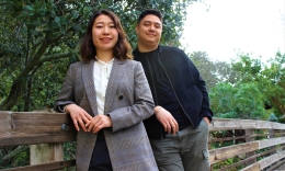Photo of smiling MBA students Miho Nakayama (left) in gray suit jacket and Dakota Flick in dark sweater behind her leaning against wooden railing of foot bridge on campus