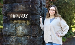 Photo of Oxford-bound student Addie Partington, standing on right with left hand on hip and smiling wearing cream colored sweater with right hand on rock column next to LIBRARY sign on the left
