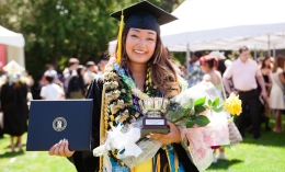 Photo of smiling graduate Sachie Ohara wearing black cap and gown and lei around her neck while holding diploma,. yellow rose, and Veritas Cup following Commencement