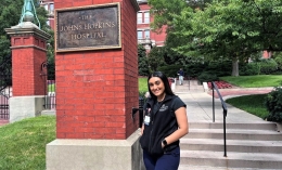 Photo of nursing alumna Tulsi Patel '22 standing and posing wearing dark vest in front of brick column sign for Johns Hopkins University with stairs leading to a building behind her