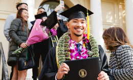 A young man wearing a black cap and gown at his graduation smiles at the camera.