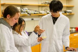 Three students in a classroom wearing lab coats analyzing light yellow substance a beaker. 