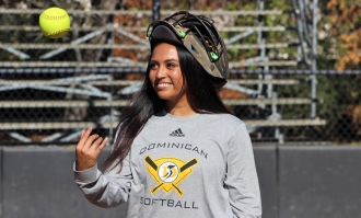 Military veteran dependent Mady Salonga '23 wearing a catcher's mask on her head tosses a yellow softball in the air near home plate in front of bleachers at Penguin Field