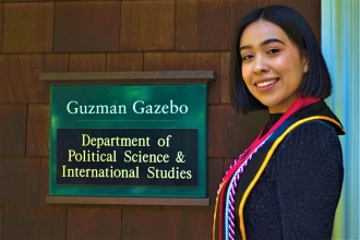 Photo of Aisleen Renteria '21 turning to face camera standing in front of brown Guzman Gazebo building with green Political Science sign