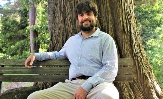 Photo of bearded Dominic Supple in light dress shirt sitting with right arm propped on bench in front of redwood tree on campus