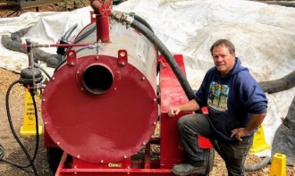 Vernon Huffman, research nursery manager at Dominican for NORS-DUC, poses in blue hoodie with right leg propped up on bumper of red steaming machine