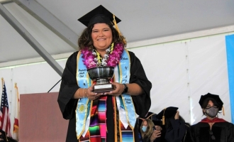Jessica Alcala '22 smiles and poses holding Veritas Cup award she received on stage in her cap and gown at Commencement 2022