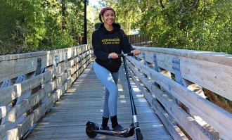 Photo of double major Autumn Jordan in black hoodie standing and posing on her scooter in middle of wooden foot bridge in John Allen Athletics Complex