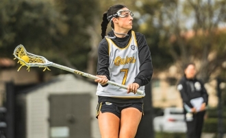Photo of Dominican women's lacrosse player Taylor Pieri, wearing white number 7 jersey, looking downfield left while holding her lacrosse stick with both hands angled to her right.