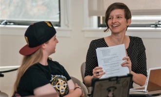 Photo of professor Katie Lewis (right) in classroom smiling and showing a document to a student