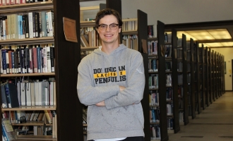Photo of Mitchell Sanders, with arms folded over gray Dominican lacrosse hoodie, standing and smiling against a book case in library with numerous book case lined up over his left shoulder in the background