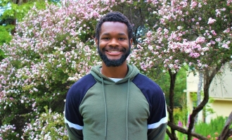 Photo of student-athlete Wakilli Bennett, standing and smiling and wearing green hoodie with blue with hands clasped behind his back in front of a bush with pink blossoms