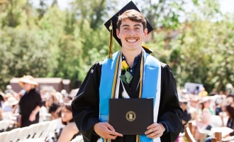 Photo of Jared Wright standing and smiling and posing for Commencement photo wearing cap and gown while holding diploma