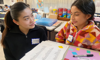 Dominican student listens to a young girl sitting at her desk at an elementary school classroom.