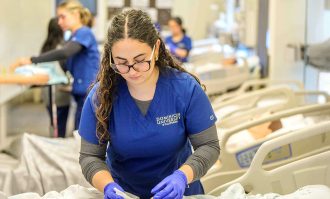 A nursing student in blue scrubs and purple gloves in the nursing simulation lab at Dominican University of California. 