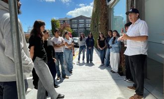 Service-learning students from Dominican University of California gather outside and listen to a man speaking.