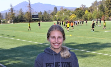 Photo of Abbie Gould in dark gray Dominican soccer hoodie standing at Kennelly Field with men's soccer practice and Mt. Tam in background 