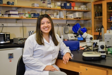 Photo of chemistry major and pre-med student Gisselle Meija '24 sitting in class laboratory wearing white lab coat with left arm propped up on lab table