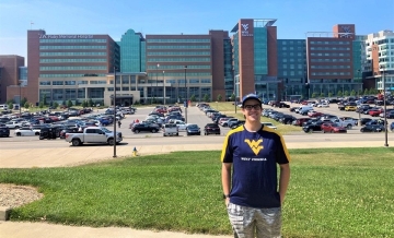 Photo of Enrique Carrera standing with hands in pockets wearing cap and blue West Virginia shirt across parking lot from WVU Hospital buildings