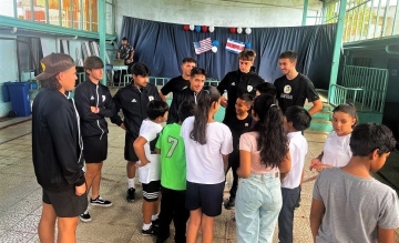 Photo of members of Dominican's men's soccer team in black tshirts and warmups engaged in conversation with elementary school students in Costa Rica in a multi purpose room