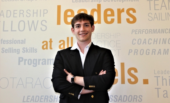 Photo of business student Augustus Muse standing with arms folded in dark sport coat with keywords on light wall behind him