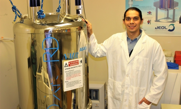 Photo of Dominican adjunct professor Joseph Morris '19 '21 MS standing on right wearing white lab coat with his right hand on placed on top of silver NMR spectrometer