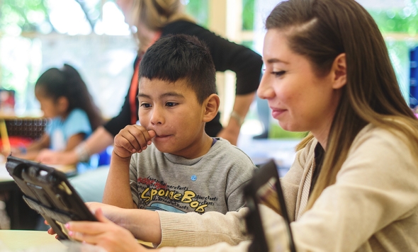Photo of a Teachers of Color scholarship recipient (right) sitting and looking into an iPad with a young student