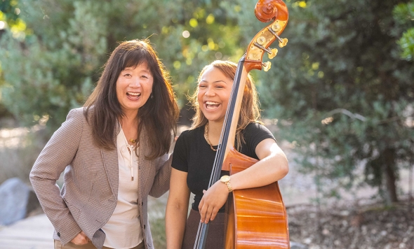 Dominican University of California Chair of Music Department, June Oh and a female student holding a bass are outdoors on the Dominican campus. Both women are laughing.