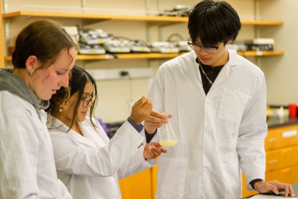 Three students in a classroom wearing lab coats analyzing light yellow substance a beaker. 