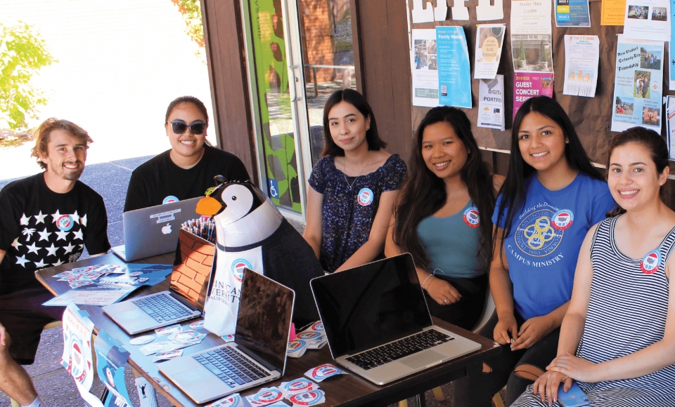 A group of Dominican University of California students sit around a table outside the Caleruega Dining Hall. Laptops are set on the table ready for students to cast their vote. A stuffed toy penguin is set in the middle of the table as decoration.