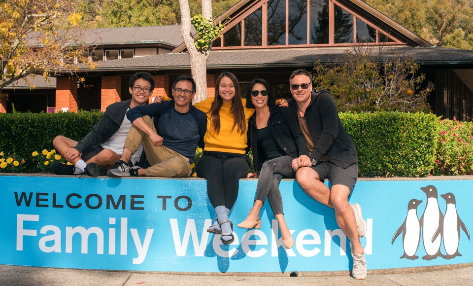 A Dominican University student family posing with the Welcome to Family Weekend banner outside Caleruega Dining Hall.