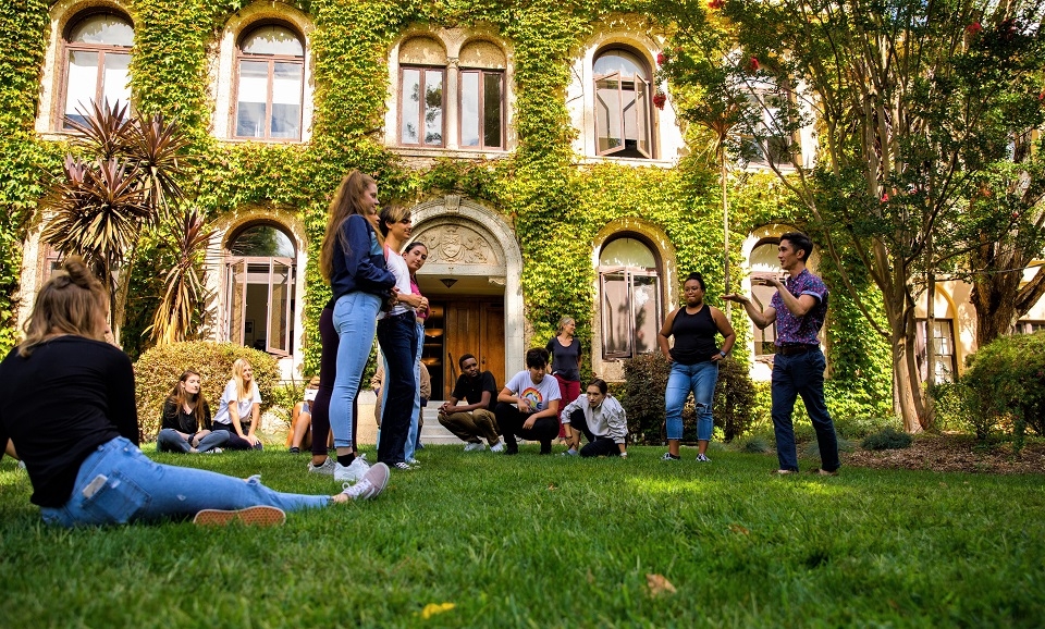 Photo of Dominican students sitting and standing around faculty member teaching on lawn outside Guzman Hall and its ivy covered walls