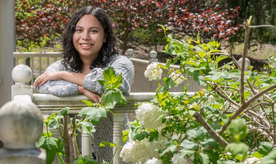 Photo of Jacquelyn Torres standing with arms folded on a porch railing with shrubs/flowers around her