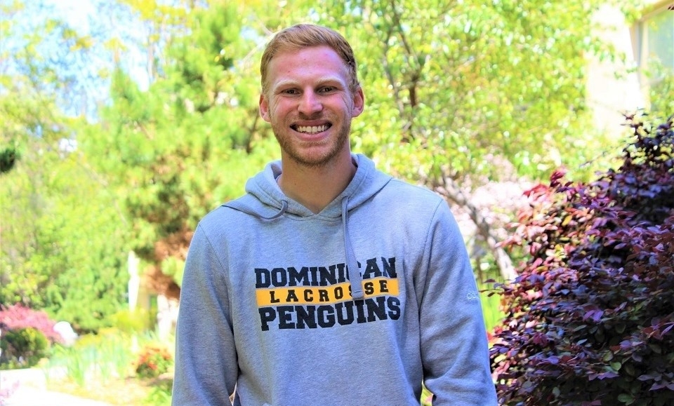 Photo of Justin Fox in gray Dominican lacrosse hoodie standing and smiling with light green tree in background