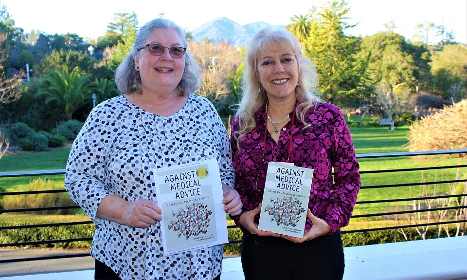 Photo of Dr. Ellen Christiansen (left) in light colored top and Dr. Luanne Linnard-Palmer in purple top posing on Meadowlands Hall porch with copy of Against Medical Advice book in their hands