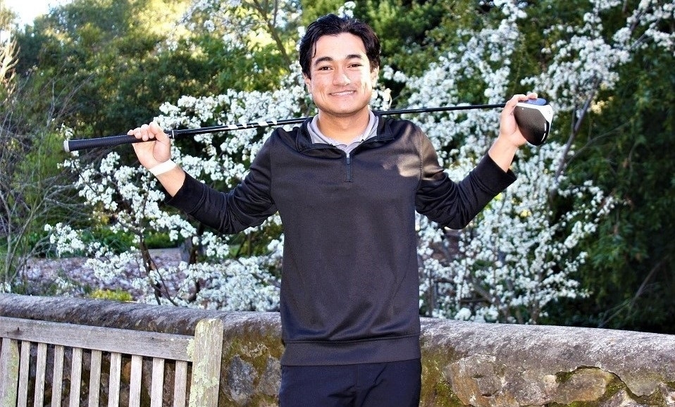 Photo of student-athlete Christian Schrodt, smiling and wearing black pants and long-sleeved shirt, posing in front of white blossomed scrub and rock wall and wooden bench while holding golf club behind his neck