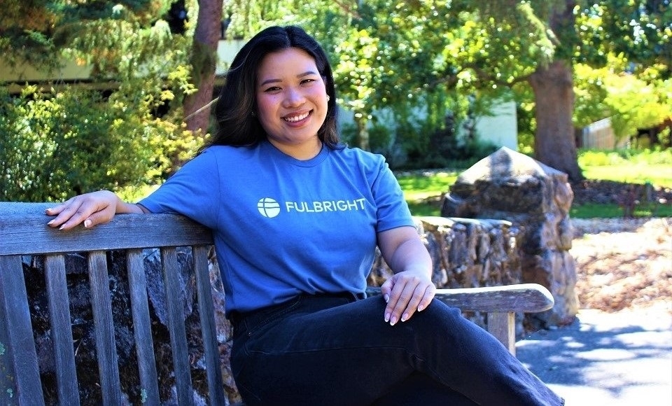 Christina Pathoumthong, wearing blue Fulbright t-shirt, sits on gray wooden bench on stone rock bridge on Dominican campus